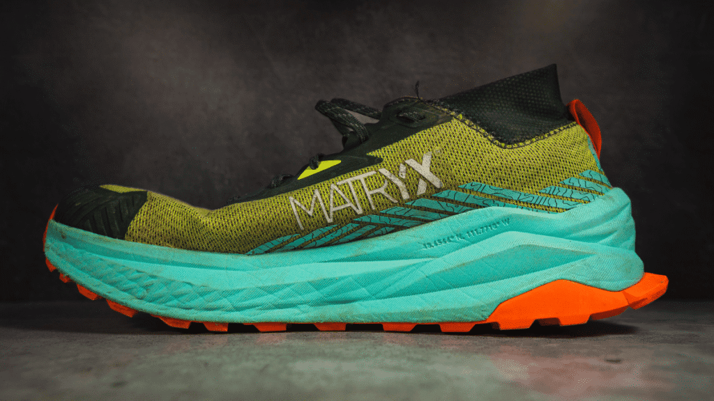 Side profile of the Altra Olympus 275 trail shoe showing the MATRYX branding on the olive green mesh upper, paired with a thick teal midsole and orange Vibram outsole, photographed against a concrete backdrop.