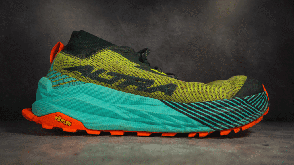 Right-side profile of the Altra Olympus 275 trail runner highlighting the bold black “Altra” logo across the green upper, with striped forefoot detail and vibrant orange and teal Vibram sole for rugged trail traction.