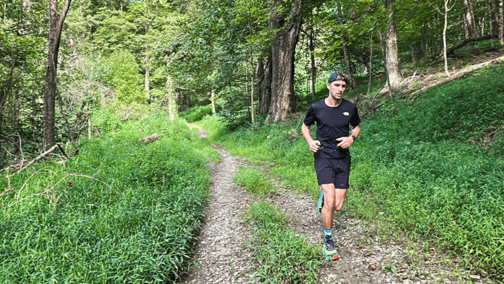Trail runner in a black outfit wearing Altra Olympus 275 shoes runs along a rocky, wooded path surrounded by dense green foliage. The sunlight filters through tall trees, highlighting the natural terrain and emphasizing the rugged trail environment.