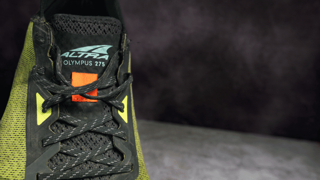 View of the Altra Olympus 2.75 tongue and lacing system, with the logo printed on the tongue and a small orange pull tab detail above black mesh.