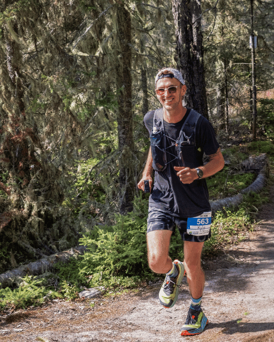 A trail runner wearing a race bib numbered 563 smiles mid-stride on a forested path. He sports a hydration vest, sunglasses, and the same Altra Olympus 275 shoes, accented by colorful socks and a backwards cap, surrounded by tall trees and dappled sunlight.
