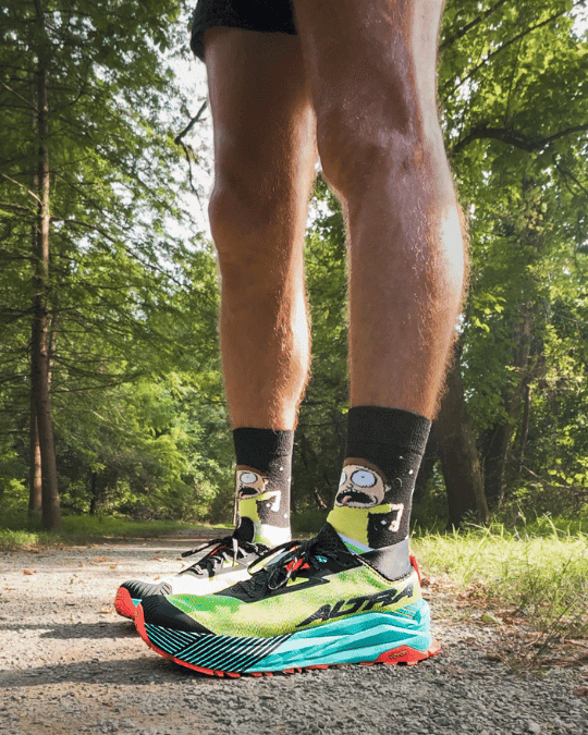 A person stands on a wooded trail wearing bright green and blue Altra Olympus 2.75 trail running shoes with red soles. Their black socks feature cartoon images of Morty from Rick and Morty with a panicked expression, adding a playful contrast to the outdoor setting.