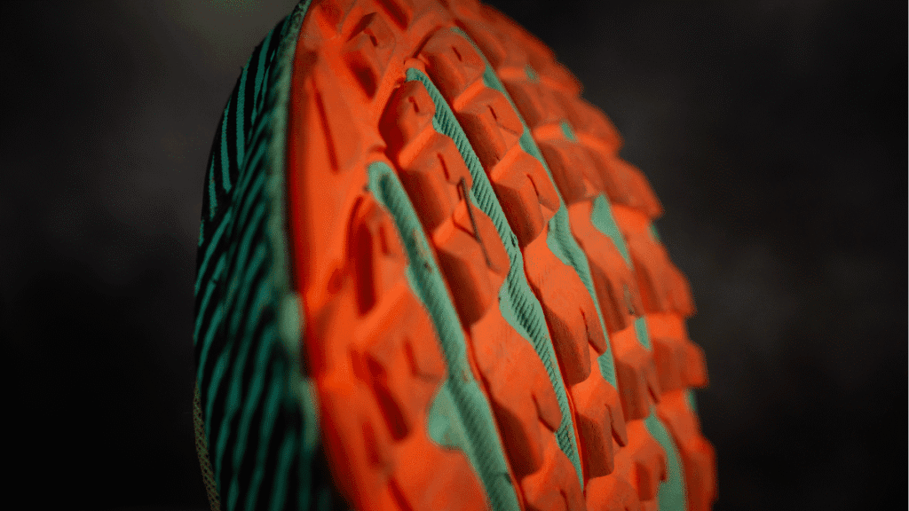 Macro shot of the shoe's aggressive orange and teal tread, emphasizing multidirectional lugs designed for trail grip and traction.