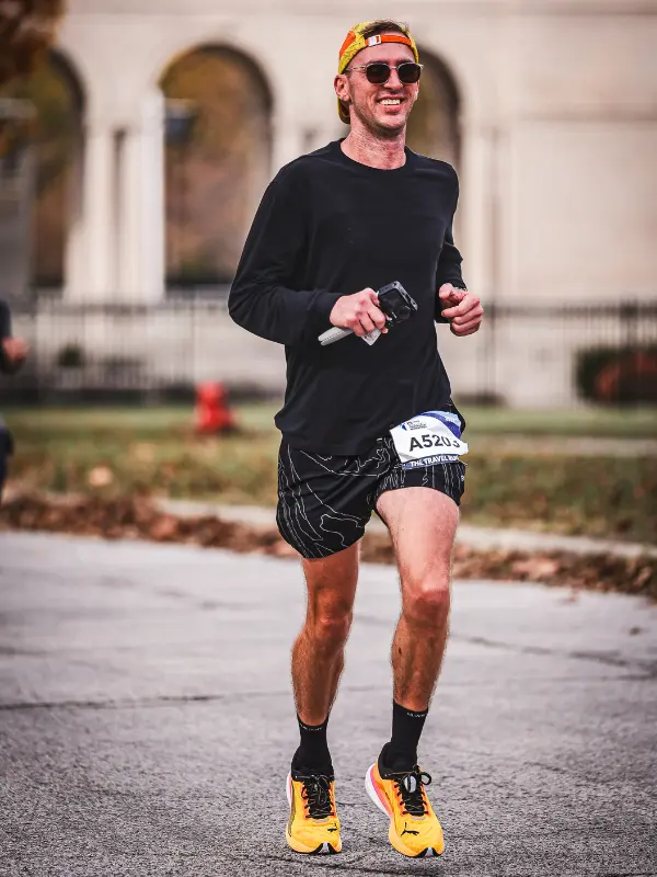 A runner in sunglasses, a backwards orange cap, and black long-sleeve top jogs during a race, holding a small camera and wearing bib number A5203.