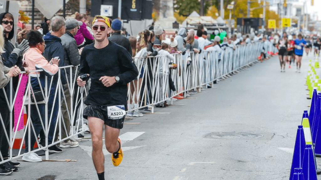 A marathon runner wearing a black long-sleeve shirt, race shorts, sunglasses, and a backwards cap nears the finish line, with cheering spectators lined up behind barricades. He holds a water bottle and sports a race bib labeled A5203, highlighting essential gear from a marathon packing list in action.