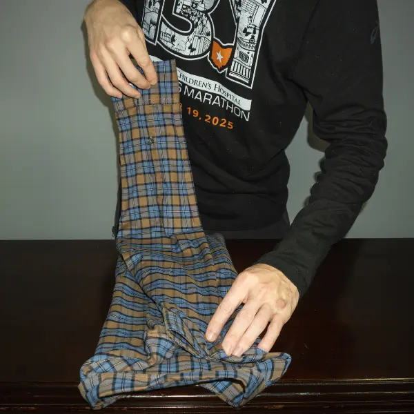 Folding the sleeves of a plaid shirt inward to create a compact rectangle before rolling or folding for travel.