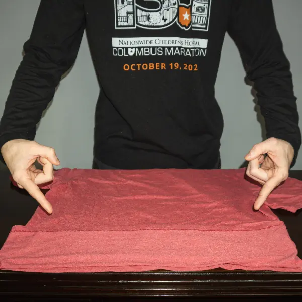 Person folding the bottom hem of a red shirt upward to start the tuck-and-roll technique for efficient travel folding.