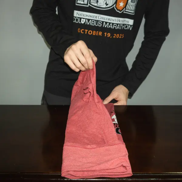 Folding one side of a red t-shirt inward after laying it flat, part of a method to minimize wrinkles and save suitcase space.