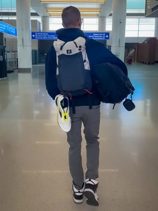 A man walks through the airport carrying two bags and a pair of running shoes clipped to his backpack. A visual reminder of the importance of planning ahead for carry-on essentials when deciding when to start packing for a trip.