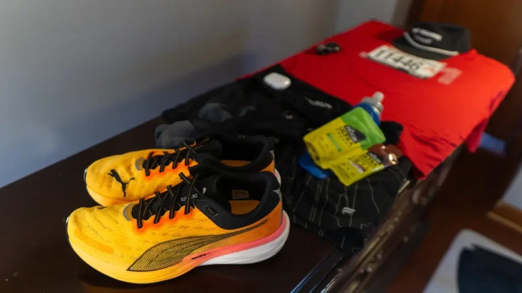 Flat lay of race day essentials on a dresser, including yellow Puma running shoes, red bib shirt, shorts, gels, and water bottle. A well-organized layout like this can help guide when to start packing for a trip, especially for athletic events.