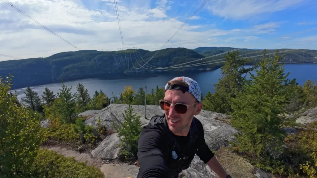 A runner takes a selfie from a rocky overlook above a wide river with forested hills stretching into the distance. Power lines cross the sky above the valley, and evergreen trees frame the viewpoint on a clear day. The image captures a scenic moment from a race route with significant elevation and expansive views.