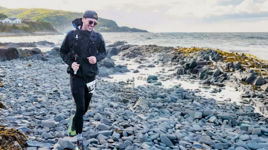 A runner in race gear runs along a rocky coastline with waves breaking against the shore nearby. The uneven stones and exposed shoreline highlight the technical terrain of the course. The image shows a challenging coastal section of a race completed in 2025.