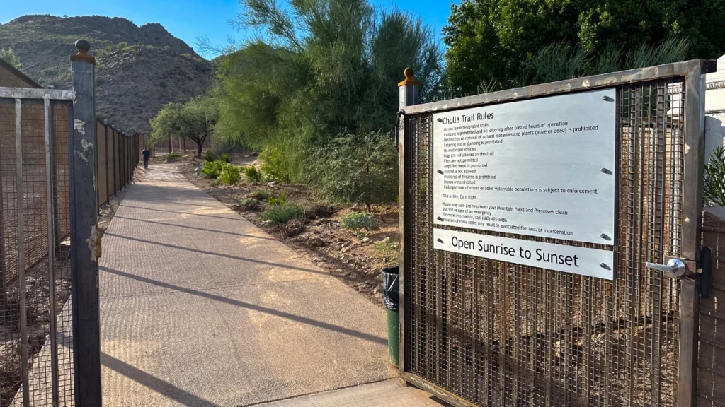 Entrance to Camelback Mountain trail with a paved path leading between desert plants and fencing. A sign reads "Cholla Trail Rules" with guidelines and "Open Sunrise to Sunset" posted at the gate.