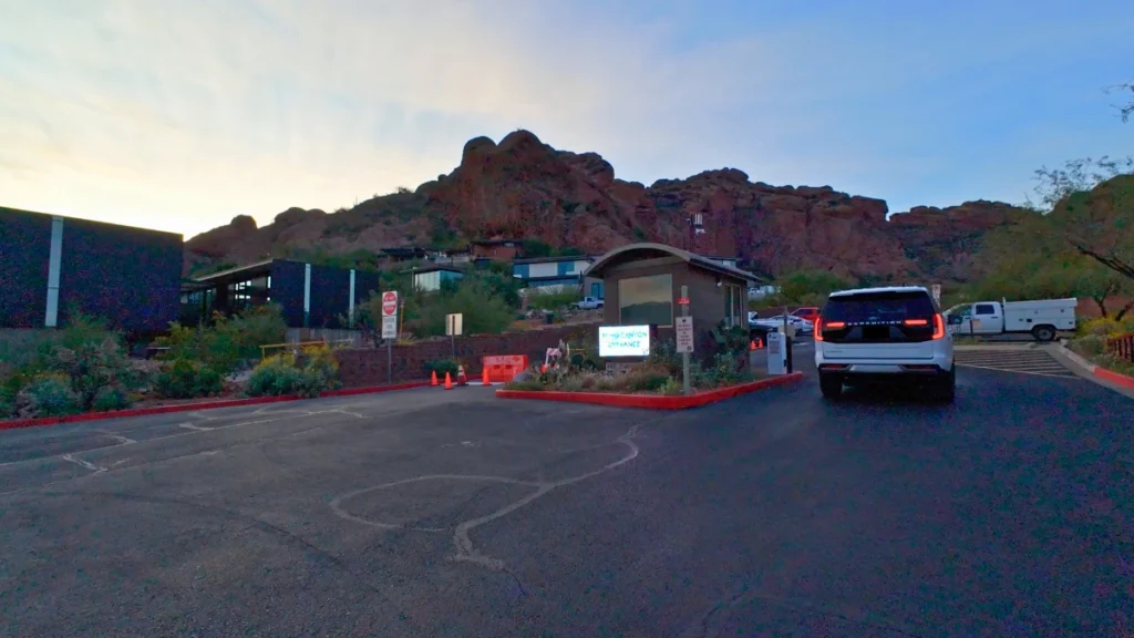 A paved, nearly full parking lot at the base of a towering red rock mountain under a bright blue sky.