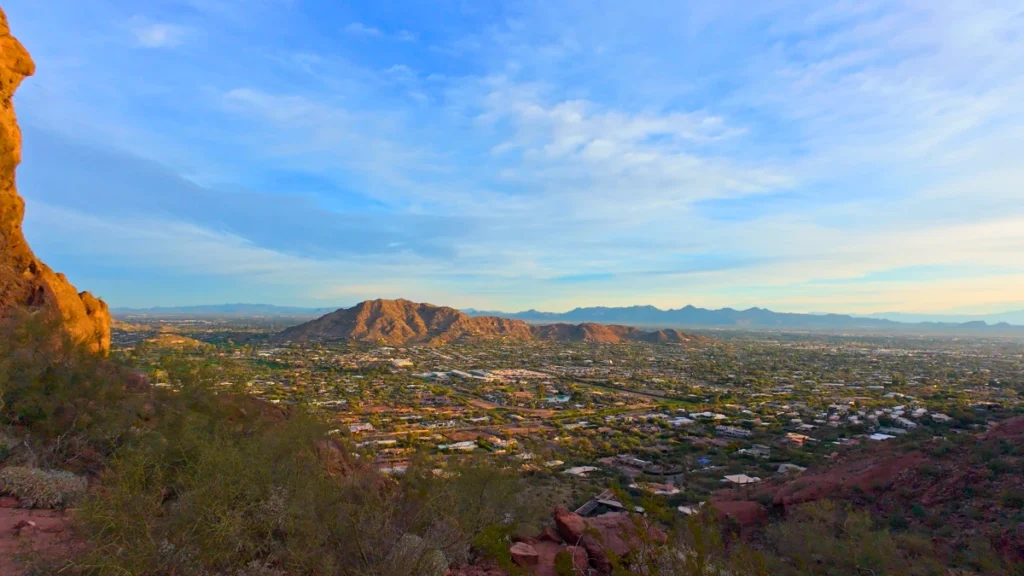 Looking out from a rocky cliff toward a jagged mountain ridge with desert scrub, some houses, and a clear horizon.