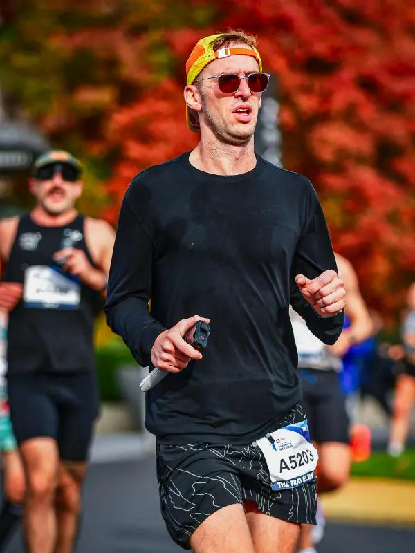 Kyle Cash running mid-race during the Indianapolis Monumental Marathon with vibrant red autumn foliage in the background, wearing black long sleeve and The Travel Runner race bib