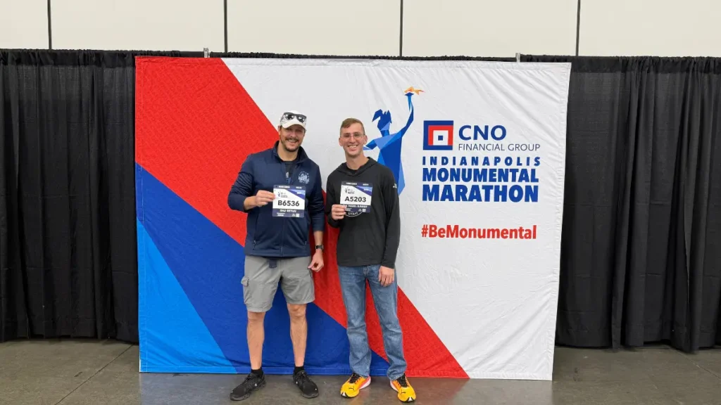 Two runners displaying their race bibs at the Indianapolis Monumental Marathon expo in front of the CNO Financial Group event banner