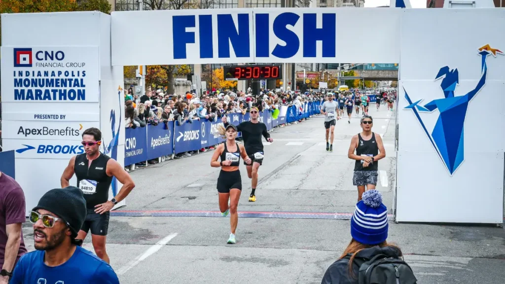 Runners crossing the finish line of the CNO Financial Group Indianapolis Monumental Marathon with the clock reading 3:30:30 and spectators packed along both sides of the course