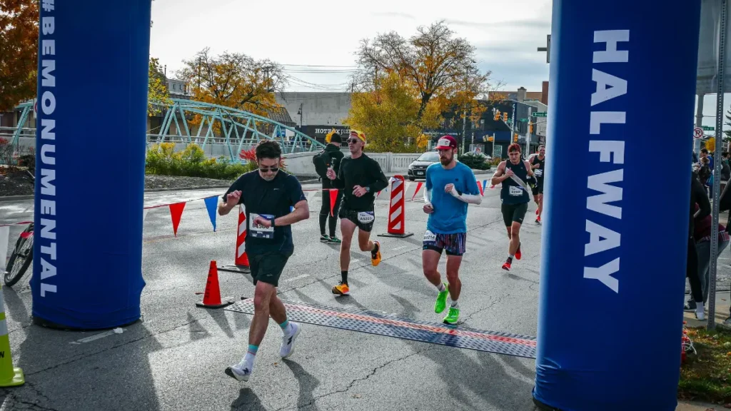 Kyle Cash crossing the halfway point arch at the Indianapolis Monumental Marathon, with fall foliage and a bridge visible in the background and the #BeMonumental banner overhead