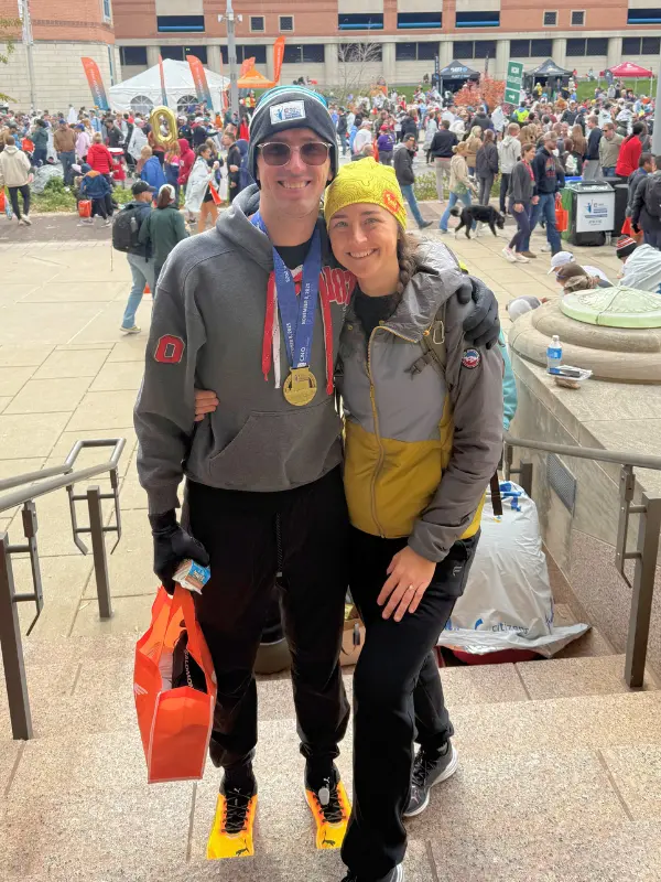 Kyle Cash and Kaitlyn posing together after the Indianapolis Monumental Marathon, Kyle wearing his finisher medal and yellow Puma racing shoes with the post-race festival crowd in the background