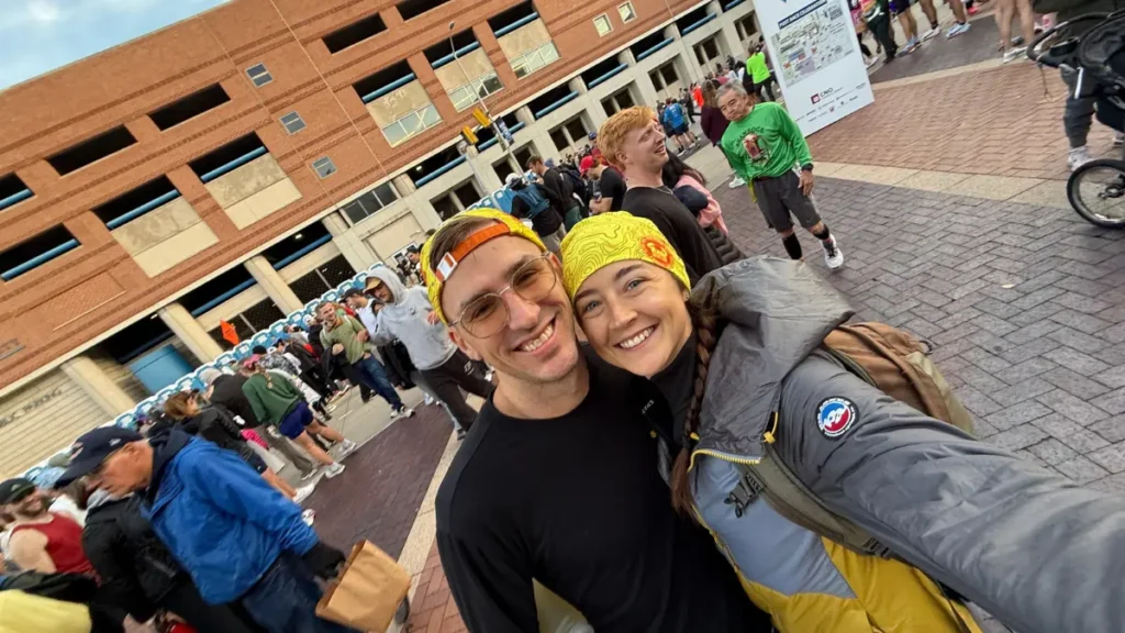 Kyle Cash and Kaitlyn smiling in a pre-race selfie at the Indianapolis Monumental Marathon start area, both wearing matching yellow headbands with hundreds of runners in the background