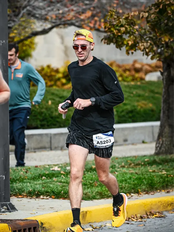 Kyle Cash in mid-stride during the Indianapolis Monumental Marathon, passing through a tree-lined city block in black long sleeve and patterned shorts