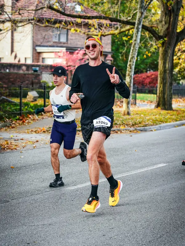 Kyle Cash flashing a peace sign and smiling through a residential neighborhood stretch of the Indianapolis Monumental Marathon, with fall color trees lining the course