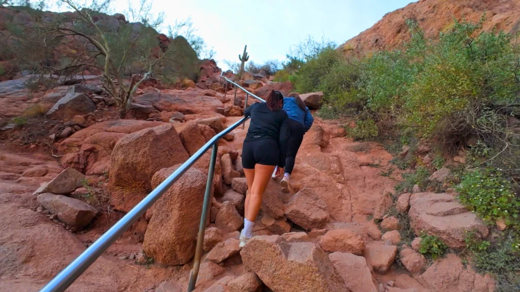 A close-up view of two women using a metal handrail bolted into red sandstone, helping climbers ascend a near-vertical pitch.