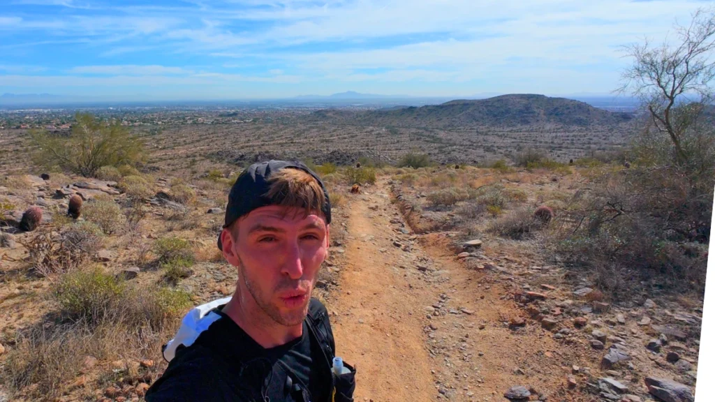 A selfie of a man in a black cap on a wide, dusty desert trail that stretches out toward a flat valley under a bright, hazy sky.