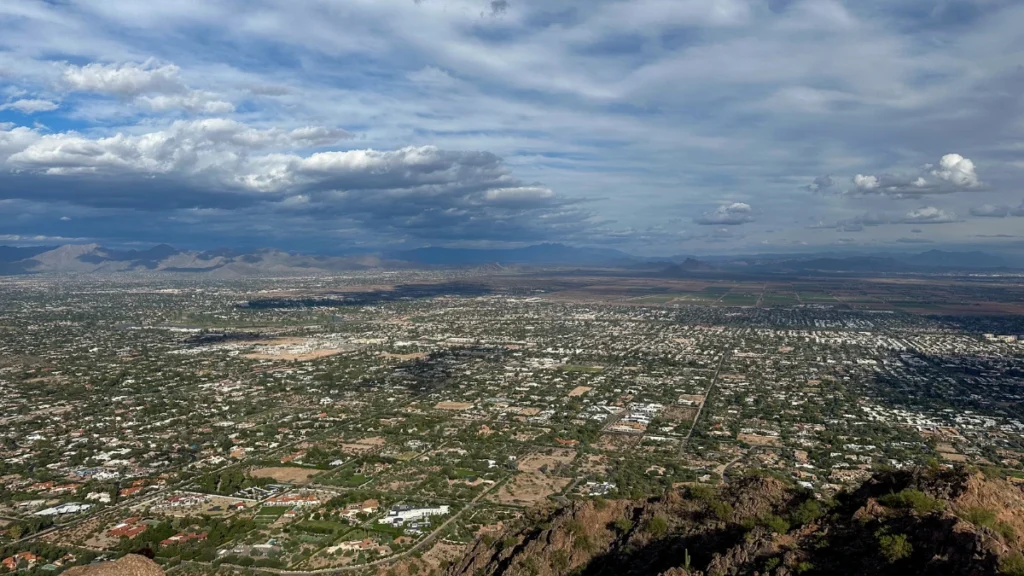 Panoramic view from Camelback Mountain overlooking a wide cityscape with scattered homes and streets under a dramatic cloudy sky. Mountains stretch across the horizon beyond the urban area.