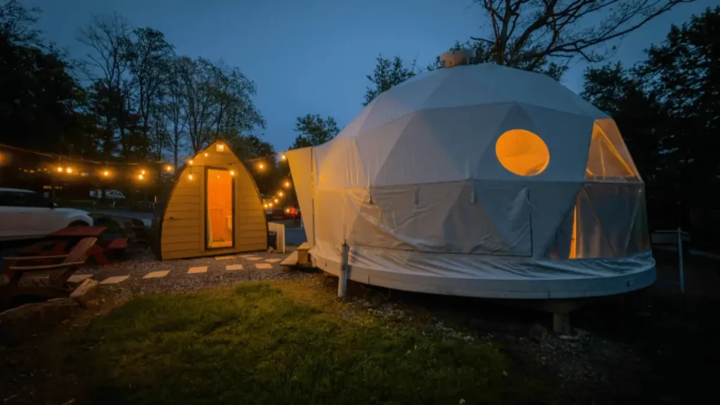 An illuminated white glamping dome yurt next to a wooden cabin pod lit by string lights at night at Bellefonte Campground in Bellefonte, PA.
