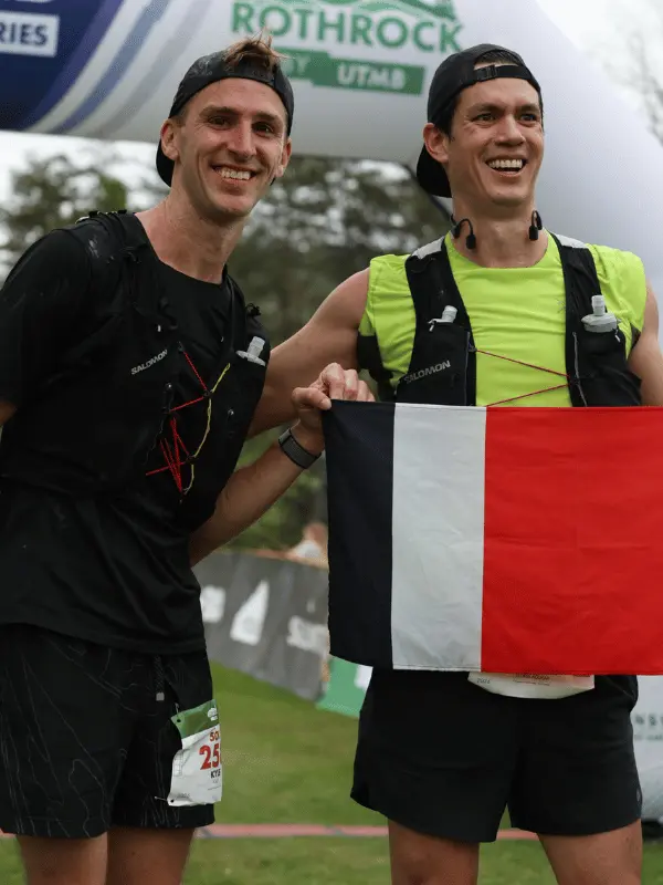Two smiling male ultramarathon runners standing together and proudly holding up a finisher flag under the Rothrock by UTMB arch.
