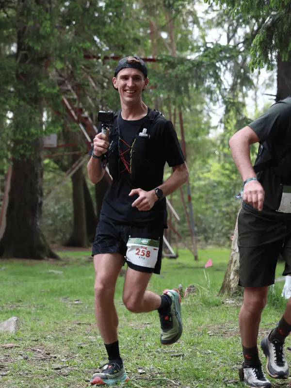 A trail runner holding a GoPro camera and wearing an hydration vest smiles while running through a grassy forest clearing.