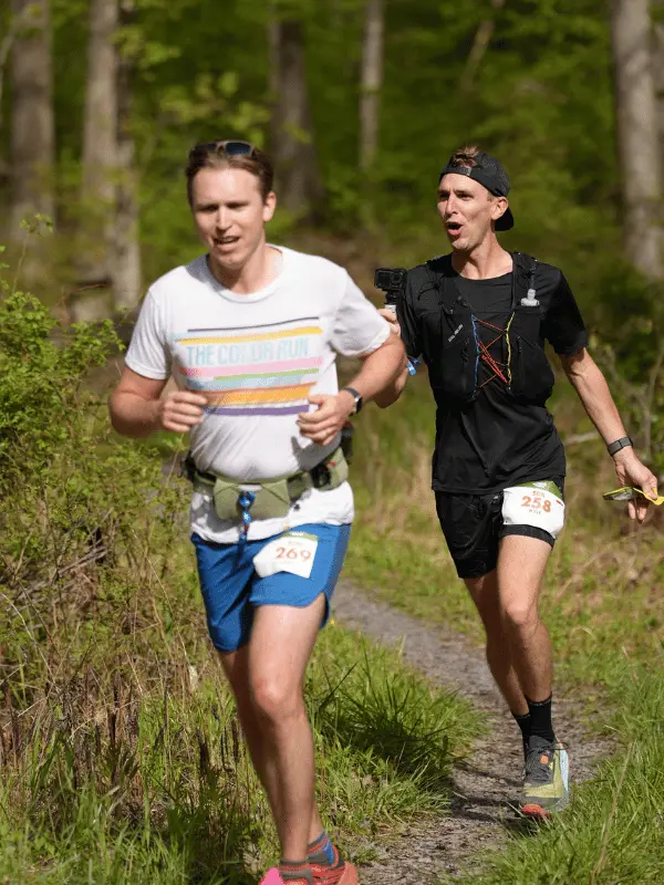 Two male runners smiling and running side-by-side on a grassy dirt trail in the woods during a sunny marathon event.