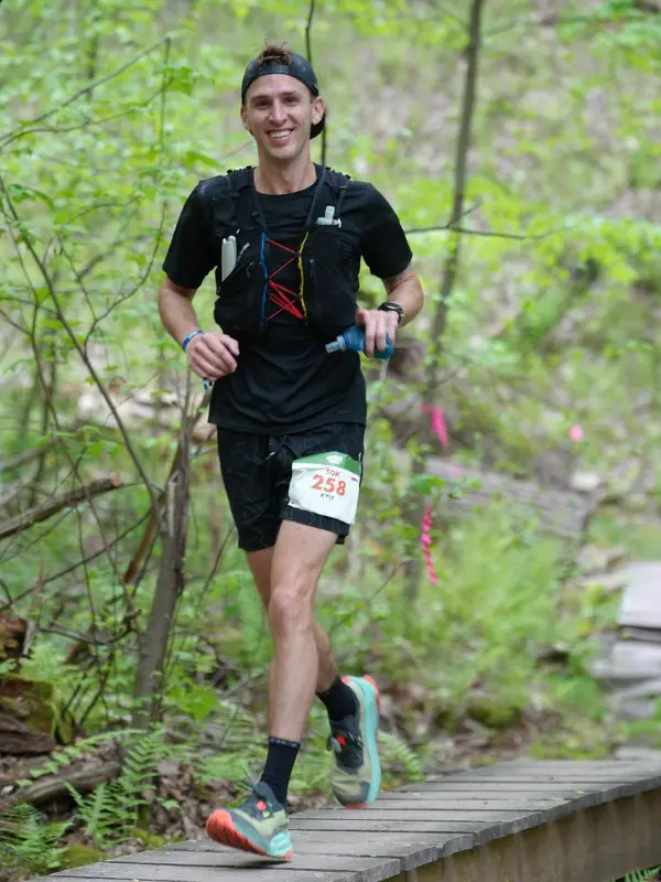 A smiling male trail runner wearing bib number 258 runs across a wooden boardwalk trail bridge in a lush green forest during the Rothrock by UTMB 50K.