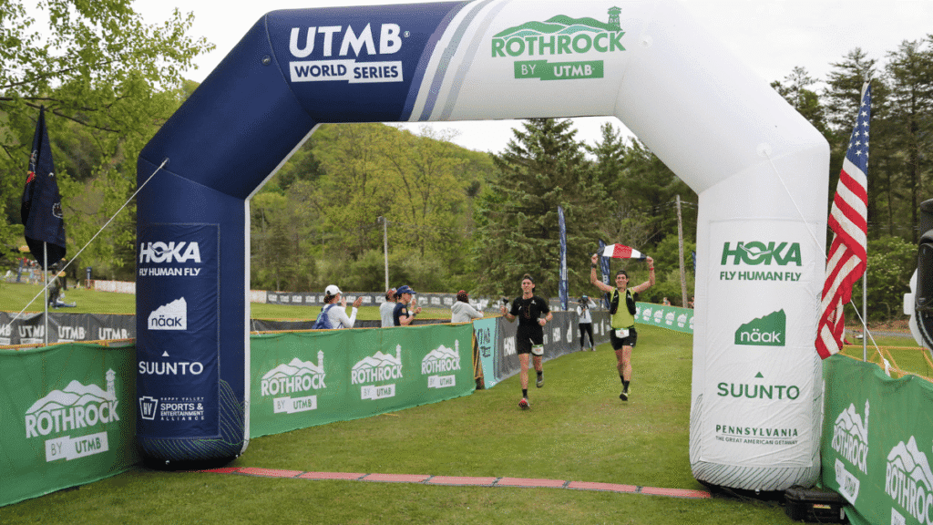 Two trail runners crossing under the large blue and white inflatable finish line arch at the Rothrock by UTMB World Series race.