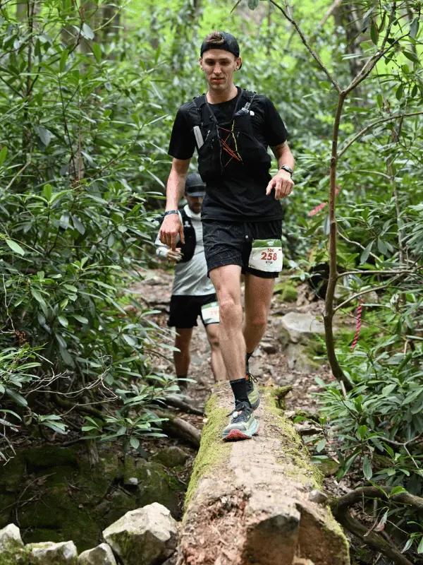 A male ultrarunner wearing bib 258 balancing across a mossy downed log bridge over a rocky creek bed during a trail race in Pennsylvania.