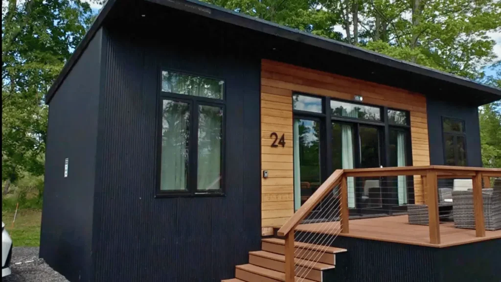 A rustic wooden tiny home cabin with a large front porch deck and string lights under a grey sky at The Hartman Center in Milroy, PA.