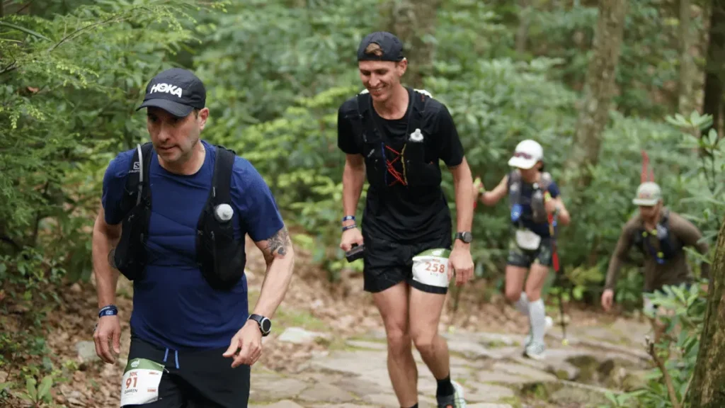 A line of ultramarathon runners power hiking up a steep, rocky stone staircase trail in a dense green forest.