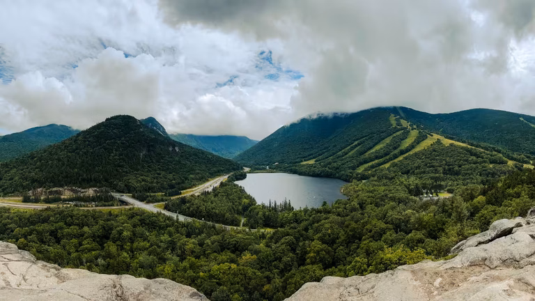 Panoramic view from Artists Bluff Trail overlooking Echo Lake, Cannon Mountain, and Franconia Notch beneath dramatic clouds.