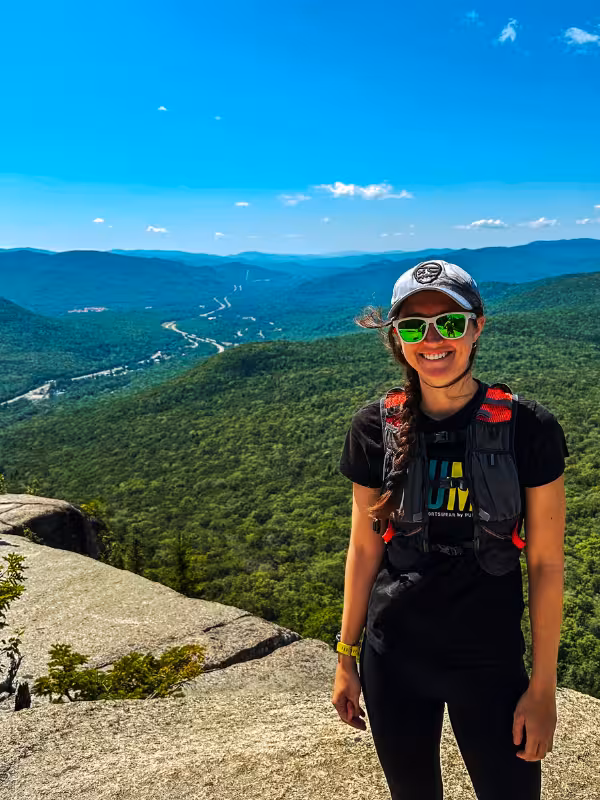 Kaitlyn standing on Mount Pemigewasset’s rocky summit with forested mountains and a winding highway behind her.