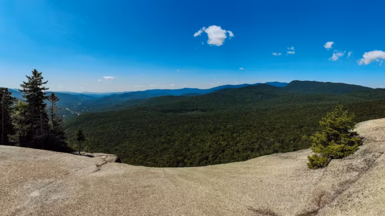 Panoramic view from Mount Pemigewasset’s broad rock ledge across a forested valley and distant mountain ridges.