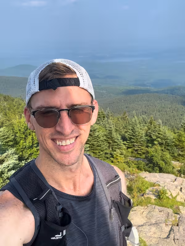 Kyle taking a selfie at the Kearsarge North summit with evergreen trees and hazy forested hills behind him.