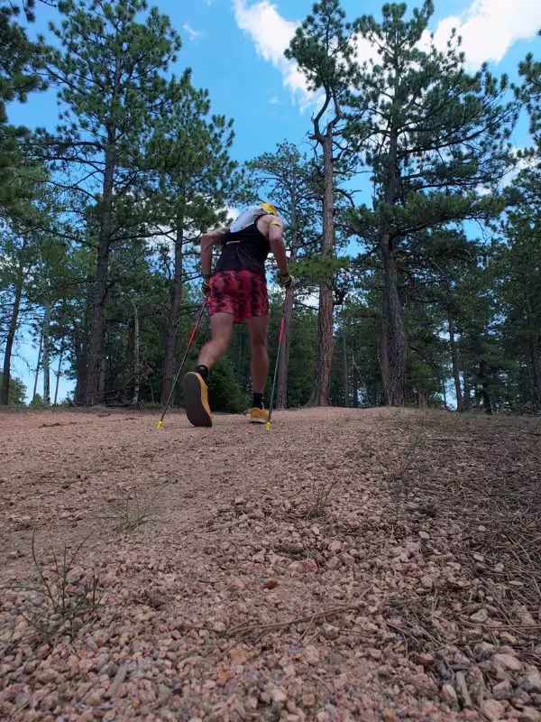 Runner using trekking poles to hike a gravelly uphill section of the Lovell Gulch Trail through pine forest.