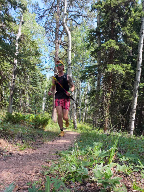 Runner carrying trekking poles on narrow dirt singletrack through aspens and evergreens on the White Gulch extension.