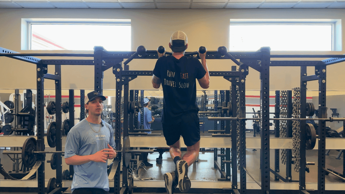 A man in a black shirt reading “Run Fast Travel Slow” performs a pull-up on a squat rack while another man in a light blue “Jim Cash” shirt and baseball cap stands nearby, speaking or giving instructions. The gym setting features weight racks, barbells, and chains in the background under bright natural light from two windows.