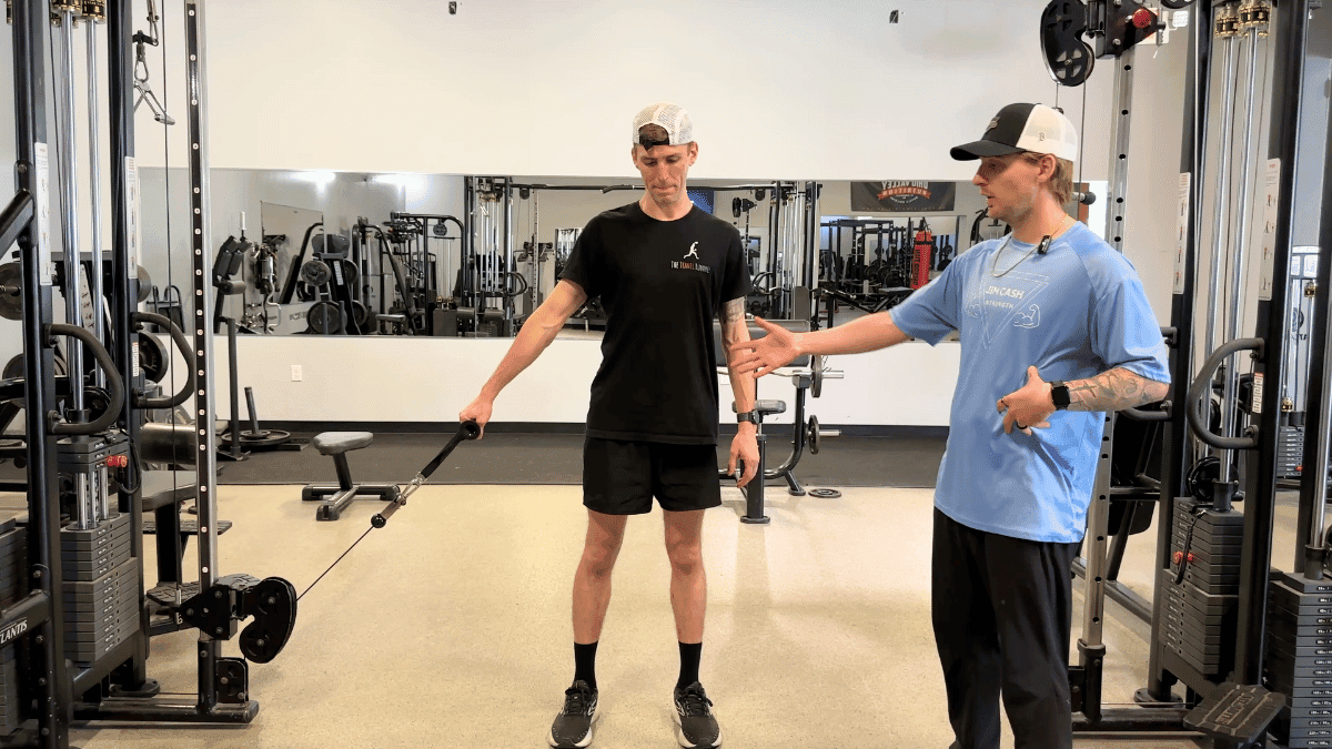Two men are in a gym, with one performing a single-arm cable lateral raise using a pulley machine while the other provides instruction. The man exercising wears a black outfit and stands upright with his arm extended to the side, while the trainer in a light blue shirt and hat gestures toward his form.